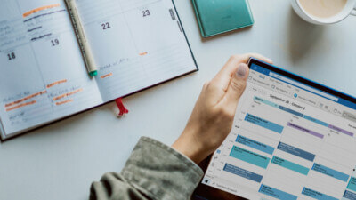 Tablet with calendar and colour-coded timeblocking is shown on a desk, with an open calendar notebook also on the table. Tasks in the book are highlighted in orange.